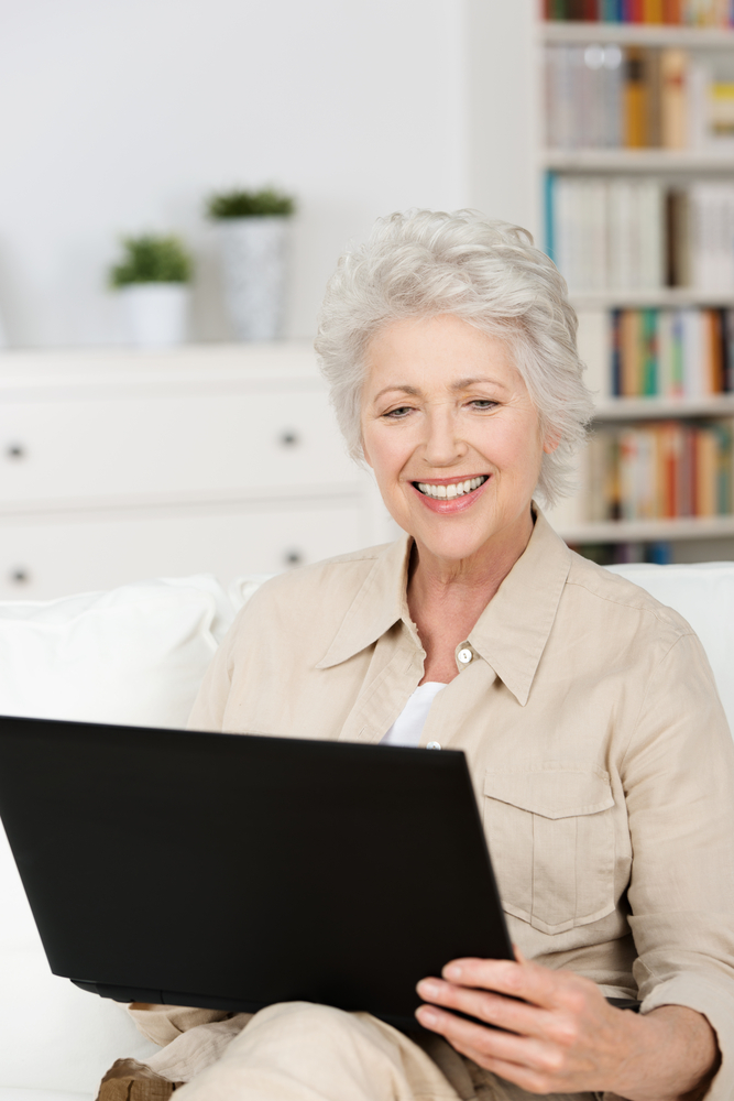 Senior woman using a laptop at home sitting on the sofa in the living room reading information on the screen with a smile